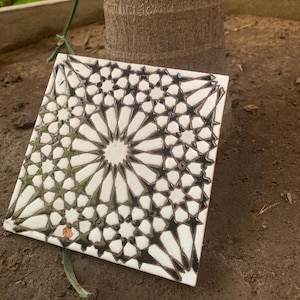 May include: A square ceramic tile featuring a complex geometric pattern in black and white. The design consists of interlocking star shapes and radiating lines that form a symmetrical, repeating motif across the entire surface of the tile.