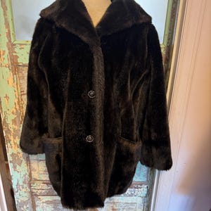 May include: A dark brown faux fur coat with a wide collar and two front pockets. The coat has two large buttons and is displayed on a wooden mannequin. The coat is a vintage style.