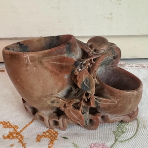 May include: A carved stone container with two open compartments and intricate floral and leaf detailing. The container is a reddish-brown color with darker accents, and sits on a patterned surface. The overall design suggests a decorative or functional item.