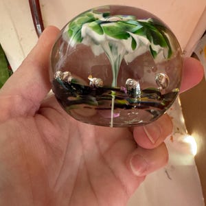May include: A clear glass paperweight with an interior design featuring white and green abstract shapes resembling a tree. The paperweight has small, clear bubbles at the base and is held in a hand.