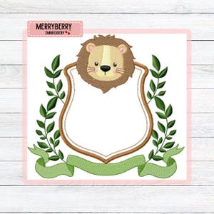 Könnte beinhalten: Gesticktes Design mit einem freundlichen Löwengesicht über einem schildförmigen Rahmen. Grüne Blätter und ein Banner umgeben den Rahmen. Das Design befindet sich auf weißem Hintergrund mit dem Text "MERRYBERRY EMBROIDERY" oben.