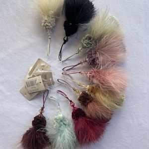 May include: A selection of decorative feather tassels in shades of white, black, pink, and burgundy. Each tassel features a beaded or knotted top and a hanging loop. Some tassels have attached tags.