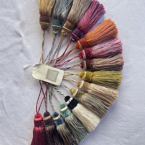 May include: A selection of decorative tassels in shades of gold, blue, pink, and red. Each tassel has a detailed design with a contrasting band and a twisted cord for hanging. A tag is attached to the tassels.