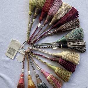 May include: A selection of decorative tassels in shades of green, burgundy, pink, and gold. Each tassel is attached to a twisted cord and a metal ring, with a tag that reads "nico decor studio".