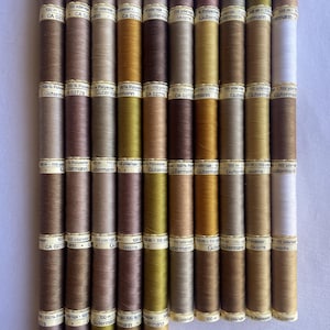 May include: A selection of sewing thread spools in a range of earthy tones, including browns, beiges, and greens. Each spool has a label with text, including "100% Polyester" and "100 m". The spools are arranged in neat vertical rows.
