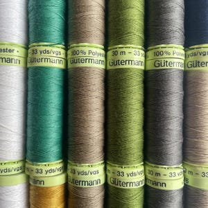 May include: A close-up shot of several Gütermann thread spools in a variety of colours, including white, green, brown, and grey. Each spool is labelled with the brand name and thread length, 30 m. The threads are tightly wound, displaying a range of neutral and earthy tones.