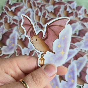 May include: A collection of bat stickers. The stickers feature a cartoon bat with brown fur and pink wings, holding a lantern. The background is a light purple with yellow stars. The stickers are on a green surface.