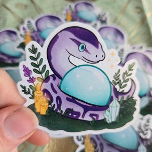 May include: A sticker featuring a purple snake with a light blue crystal ball. The snake is surrounded by green foliage, candles, and crystals. The sticker has a white border and is held by a hand.