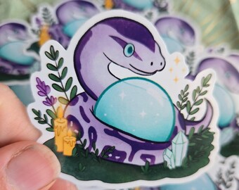 Può includere: Un adesivo raffigurante un serpente viola che tiene una sfera di cristallo azzurra. Il serpente è circondato da fogliame verde, candele e cristalli. L'adesivo ha un bordo bianco ed è tenuto da una mano.