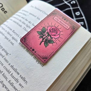 Pode incluir: Um marcador de livro rosa com uma ilustra&ccedil;&atilde;o de rosa e a inscri&ccedil;&atilde;o "the maiden". O marcador de livro est&aacute; colocado num livro aberto. A rosa tem folhas verdes e um desenho de sol.