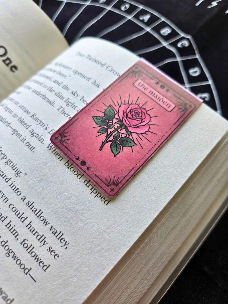 Pode incluir: Um marcador de livro rosa com uma ilustra&ccedil;&atilde;o de rosa e a inscri&ccedil;&atilde;o "the maiden". O marcador de livro est&aacute; colocado num livro aberto. A rosa tem folhas verdes e um desenho de sol.
