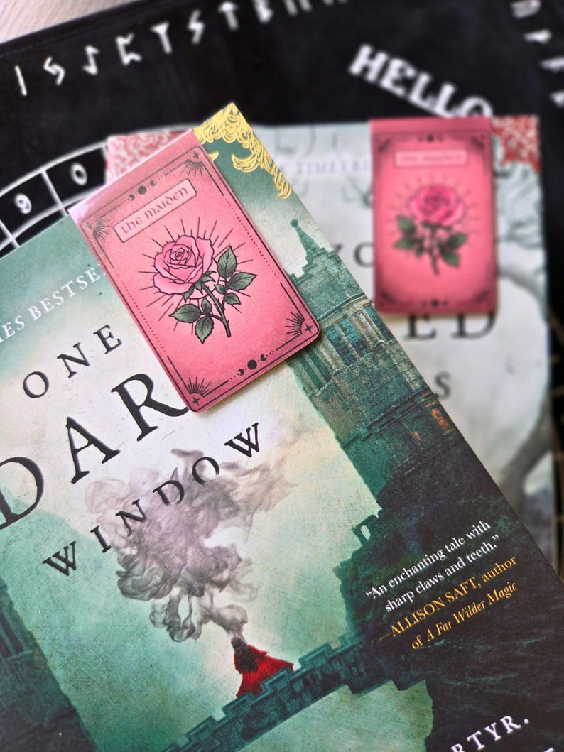 Pode incluir: Marcador de livros retangular rosa com uma ilustra&ccedil;&atilde;o de rosa e a inscri&ccedil;&atilde;o "the maiden". O marcador de livros est&aacute; colocado em cima de um livro intitulado "ONE DARK WINDOW". Outro marcador de livros &eacute; vis&iacute;vel no fundo.