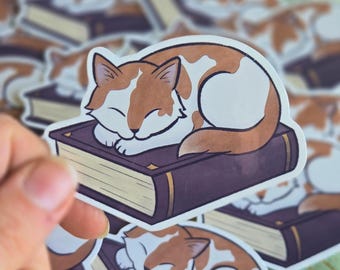 May include: A sticker featuring a sleeping cat with orange and white fur, resting on a purple book. The sticker has a white border and is held by a hand, with other similar stickers in the background.
