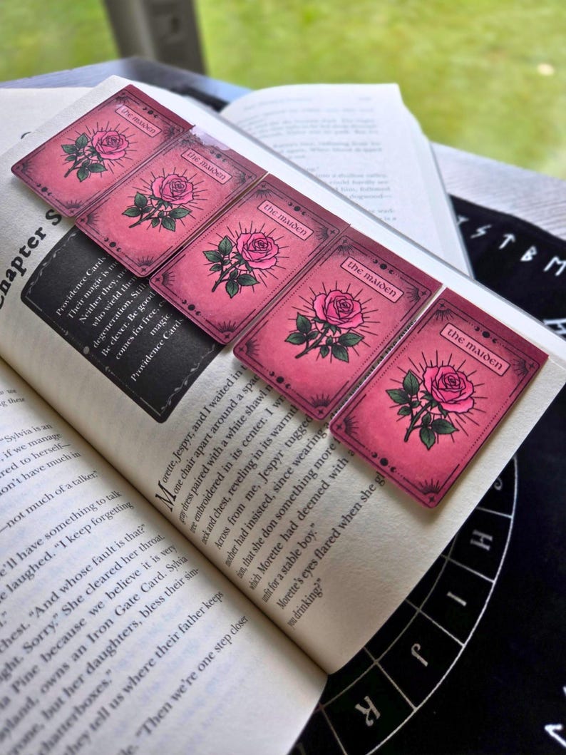 Pode incluir: Cinco marcadores de livros retangulares cor-de-rosa com uma ilustra&ccedil;&atilde;o de rosa e o texto "The Maiden" est&atilde;o colocados em um livro aberto. Os marcadores de livros t&ecirc;m uma est&eacute;tica vintage com uma borda rosa escura e uma rosa.