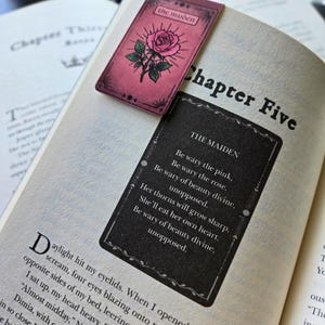 Pode incluir: Um livro aberto no Cap&iacute;tulo Cinco, com dois marcadores estilo tar&ocirc;. Um &eacute; rosa com uma ilustra&ccedil;&atilde;o de rosa e as palavras "the maiden". O outro &eacute; preto com um poema sobre "the maiden".