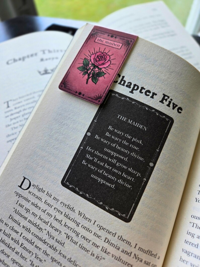 Pode incluir: Um livro aberto no Cap&iacute;tulo Cinco, com dois marcadores estilo tar&ocirc;. Um &eacute; rosa com uma ilustra&ccedil;&atilde;o de rosa e as palavras "the maiden". O outro &eacute; preto com um poema sobre "the maiden".