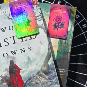 Pode incluir: Dois marcadores de livros "The Maiden" est&atilde;o em cima de livros. Um tem um acabamento hologr&aacute;fico, o outro &eacute; rosa com uma ilustra&ccedil;&atilde;o de rosa. Os livros t&ecirc;m t&iacute;tulos como "Two Twisted Crowns" e "Monster, Martyr".