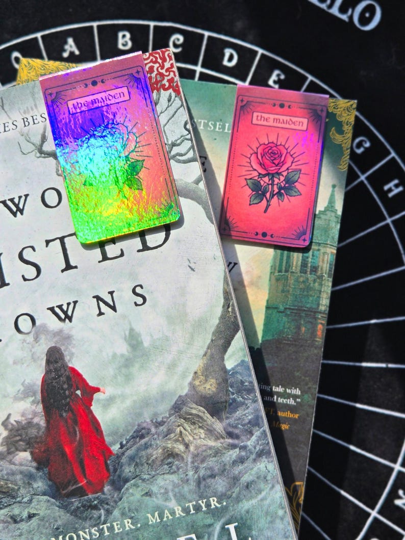 Pode incluir: Dois marcadores de livros "The Maiden" est&atilde;o em cima de livros. Um tem um acabamento hologr&aacute;fico, o outro &eacute; rosa com uma ilustra&ccedil;&atilde;o de rosa. Os livros t&ecirc;m t&iacute;tulos como "Two Twisted Crowns" e "Monster, Martyr".