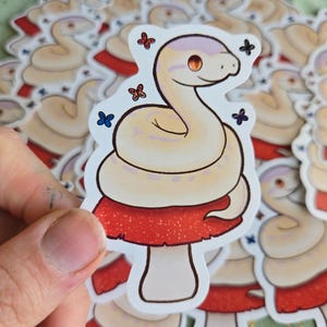 May include: A sticker featuring a cartoon snake with a cream-colored body and a light purple head, resting on a red mushroom with glitter accents. Small butterfly illustrations surround the snake. The sticker is held in a hand.