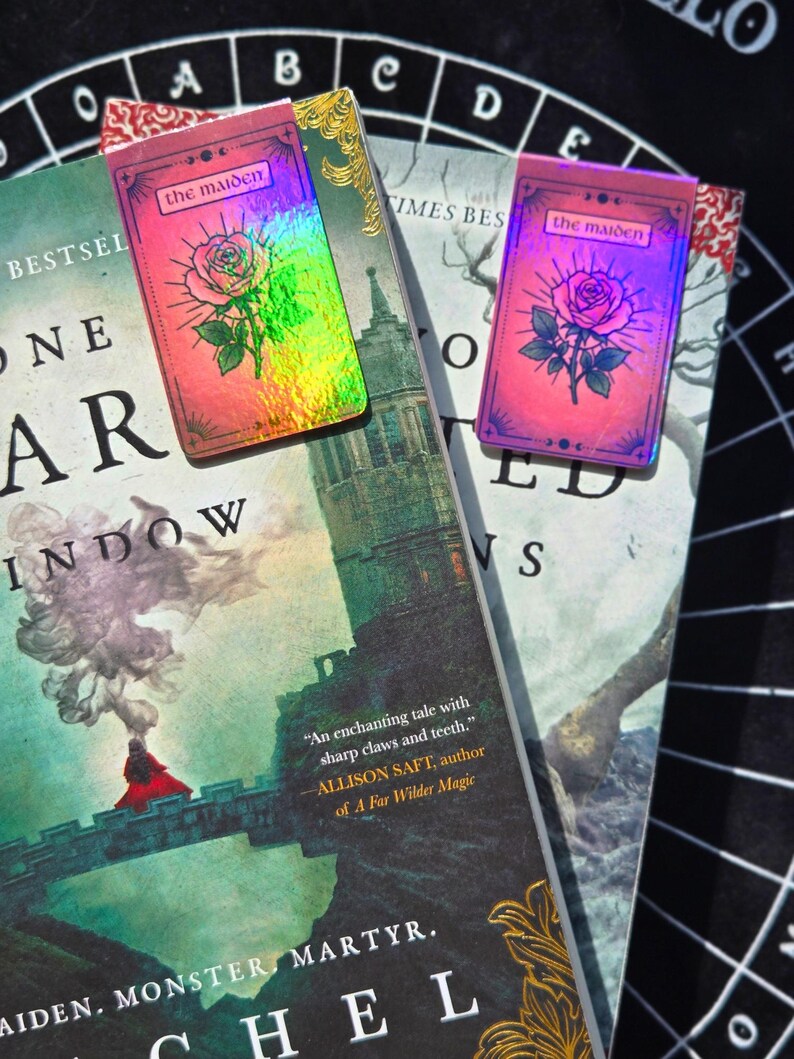 Pode incluir: Tr&ecirc;s livros com marcadores decorativos. Os marcadores apresentam uma ilustra&ccedil;&atilde;o de rosa e o texto "the maiden" em um fundo rosa. Os livros t&ecirc;m t&iacute;tulos como "One Star" e "Times Best". O fundo &eacute; uma superf&iacute;cie preta com letras brancas.