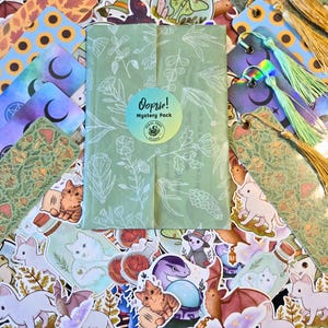 Puede incluir: Una colecci&oacute;n de pegatinas, marcadores y un paquete misterioso. Las pegatinas presentan varias criaturas de fantas&iacute;a y dise&ntilde;os bot&aacute;nicos. Los marcadores tienen detalles de borlas. El paquete misterioso est&aacute; envuelto en papel verde con ilustraciones florales y el texto "Oopsie! Mystery Pack".