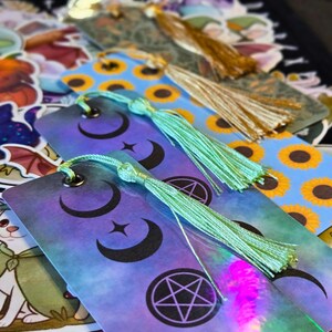 Puede incluir: Marcadores con dise&ntilde;os celestiales y borlas. Los marcadores presentan un degradado de morado y azul con lunas crecientes, estrellas y un pentagrama. Algunos tienen un patr&oacute;n de girasoles. Cada marcador tiene una borla verde menta o dorada.