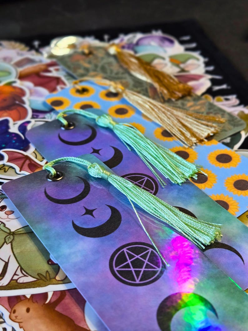 Puede incluir: Marcadores con dise&ntilde;os celestiales y borlas. Los marcadores presentan un degradado de morado y azul con lunas crecientes, estrellas y un pentagrama. Algunos tienen un patr&oacute;n de girasoles. Cada marcador tiene una borla verde menta o dorada.