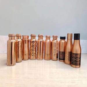 Puede incluir: Una colección de diez botellas de agua de cobre dispuestas en fila. Las botellas presentan varios acabados, incluyendo texturas lisas, martilladas y grabadas. Algunas tienen tapas de cobre, mientras que otras tienen tapas negras con patrones.