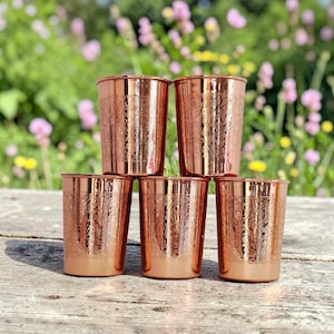 May include: A set of five cylindrical copper tumblers arranged in a pyramid formation. Each tumbler features a polished metallic finish and an intricate, embossed floral pattern covering the entire exterior surface of the vessel walls.