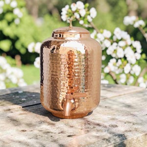 Puede incluir: Un dispensador de bebidas cilíndrico de cobre con textura martillada, una tapa a juego con un pequeño pomo central y un grifo funcional fijado en la parte inferior frontal. La superficie metálica refleja la luz sobre su cuerpo.