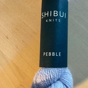 May include: A skein of Shibui Knits Pebble yarn in a light gray color. The yarn is wrapped with a black label that reads "SHIBUI KNITS" and "PEBBLE" in white text. The words "Color With Yarn" are also visible.