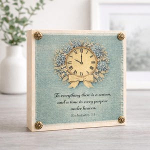 May include: Square wooden block with a light blue fabric background. A clock design with a floral wreath and bow is centered on the block. The text "To everything there is a season..." is printed below the clock. Decorative corner studs.