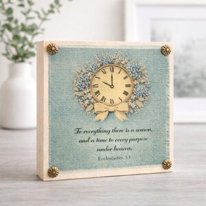May include: Square wooden block with a light blue fabric background. A clock design with a floral wreath and bow is centered on the block. The text "To everything there is a season..." is printed below the clock. Decorative corner studs.