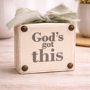May include: A decorative wooden block with the text "God's got this" in gray lettering. The block is adorned with a sage green bow and four decorative bronze-colored studs. The item is placed on a light-colored wooden surface.