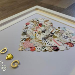 Peut inclure: Un collage en forme de cœur encadré, composé de découpes de papier à motifs. Le collage comprend des éléments floraux, rayés et imprimés de texte. Des lettres dorées épellent "DOG" et une date est en dessous. Le cadre est blanc.