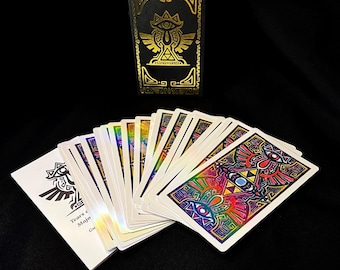 May include: A black card deck box with gold foil detailing and a matching deck of cards. The cards feature colorful, intricate designs and the text "Tears of Majo Gu". The cards are fanned out on a black surface.