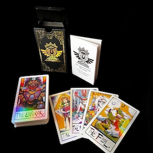 May include: A tarot card deck with a black box and a booklet. The box and booklet feature a gold emblem. Several tarot cards are visible, including "The Emperor" and "The Fool", with colorful illustrations and holographic accents. The deck is themed around The Legend of Zelda.