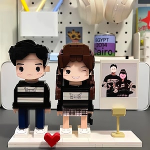 Puede incluir: Dos figuras de pareja estilo bloque, un hombre y una mujer, con camisas a rayas blancas y negras. La figura femenina guiña un ojo. También hay un corazón rojo y una foto de la pareja.