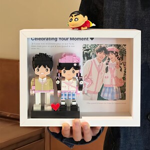 May include: A white framed photo display with a picture of a couple, two block figures, and a cartoon character. The frame has the text "Celebrating Your Moment" and a French quote. The block figures are of a couple with a red heart.