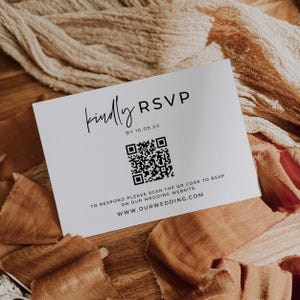 May include: A white RSVP card with black text and a QR code. The card reads "Kindly RSVP" and includes the date "BY 16.09.XX" and the website address. The card is on a wooden surface with fabric.