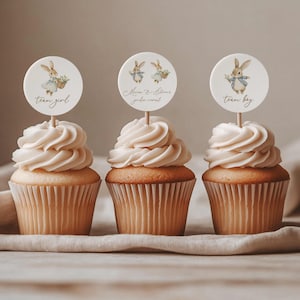 May include: Three cupcakes with white frosting and decorative toppers. The toppers feature watercolour illustrations of bunnies and text that reads "team girl", "Mom & Showers gender reveal", and "team boy". The cupcakes sit on a neutral-coloured cloth.