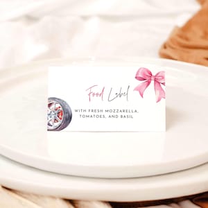 Könnte beinhalten: Eine rechteckige weiße Lebensmitteletikettenkarte mit einer rosa Aquarellschleife auf der rechten Seite und einer Illustration eines Autorads auf der linken Seite. Der Text lautet Food Label gefolgt von WITH FRESH MOZZARELLA, TOMATOES, AND BASIL.