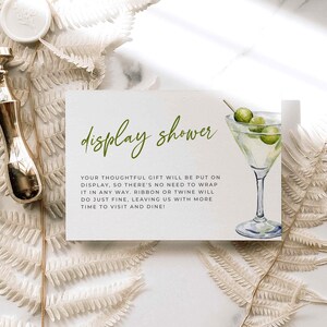 Può includere: Un cartoncino rettangolare bianco con il testo in corsivo verde display shower e un'illustrazione ad acquerello di un bicchiere da martini con tre olive. Sotto il testo, istruzioni stampate in nero spiegano le preferenze per i regali.
