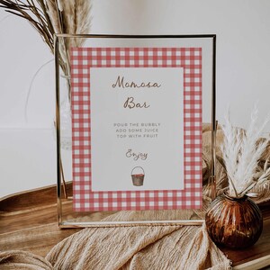 Może przedstawiać: Oprawiony znak z czerwono-białą obwódką w kratkę. Znak głosi "Mimosa Bar" w eleganckim skrypcie, z instrukcjami dodania soku i owoców. Mała ilustracja wiadra znajduje się na dole.