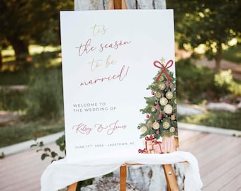May include: A white wedding welcome sign featuring the text 'tis the season to be married!' in red and gold script, followed by 'WELCOME TO THE WEDDING OF', 'Kelsey & James', and 'JUNE 17TH, 20XX, LAKETOWN, NC'. A watercolor Christmas tree illustration.