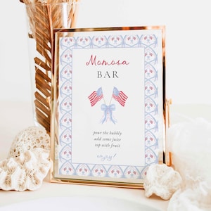 May include: A rectangular sign in a gold metal frame featuring the text Momosa BAR with two crossed American flags tied with a light blue ribbon. Below the flags, the text reads: pour the bubbly, add some juice, top with fruit, enjoy.