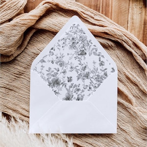 May include: A white envelope with a pointed flap featuring an interior liner printed with a detailed grey and white botanical floral pattern. The envelope is open, displaying the intricate illustration of various flowers and leaves inside the flap.