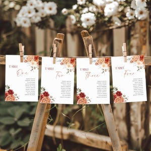Puede incluir: Cuatro tarjetas de mesa rectangulares blancas para bodas colgadas de un marco de madera con pinzas. Cada tarjeta muestra la palabra TABLE seguida de One, Two, Three o Four en cursiva, una lista de nombres e ilustraciones florales en rojo y naranja.