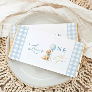 Op de afbeelding: Een rechthoekige chocoladereepverpakking met een lichtblauwe en witte gingham-rand. Het midden toont een illustratie van een gouden puppy met een blauwe strik, een blauwe ballon en de tekst Liam's ONE golden year in blauw schrift.