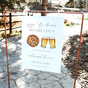 May include: A white rectangular sign hanging from a copper frame featuring the text pizza & brews BEFORE I DO'S, WELCOME TO OUR rehearsal dinner, SARAH & JAMES, JUNE 17TH 20XX - ROSEWOOD, with a watercolor illustration of a pepperoni pizza and two beer glasses.